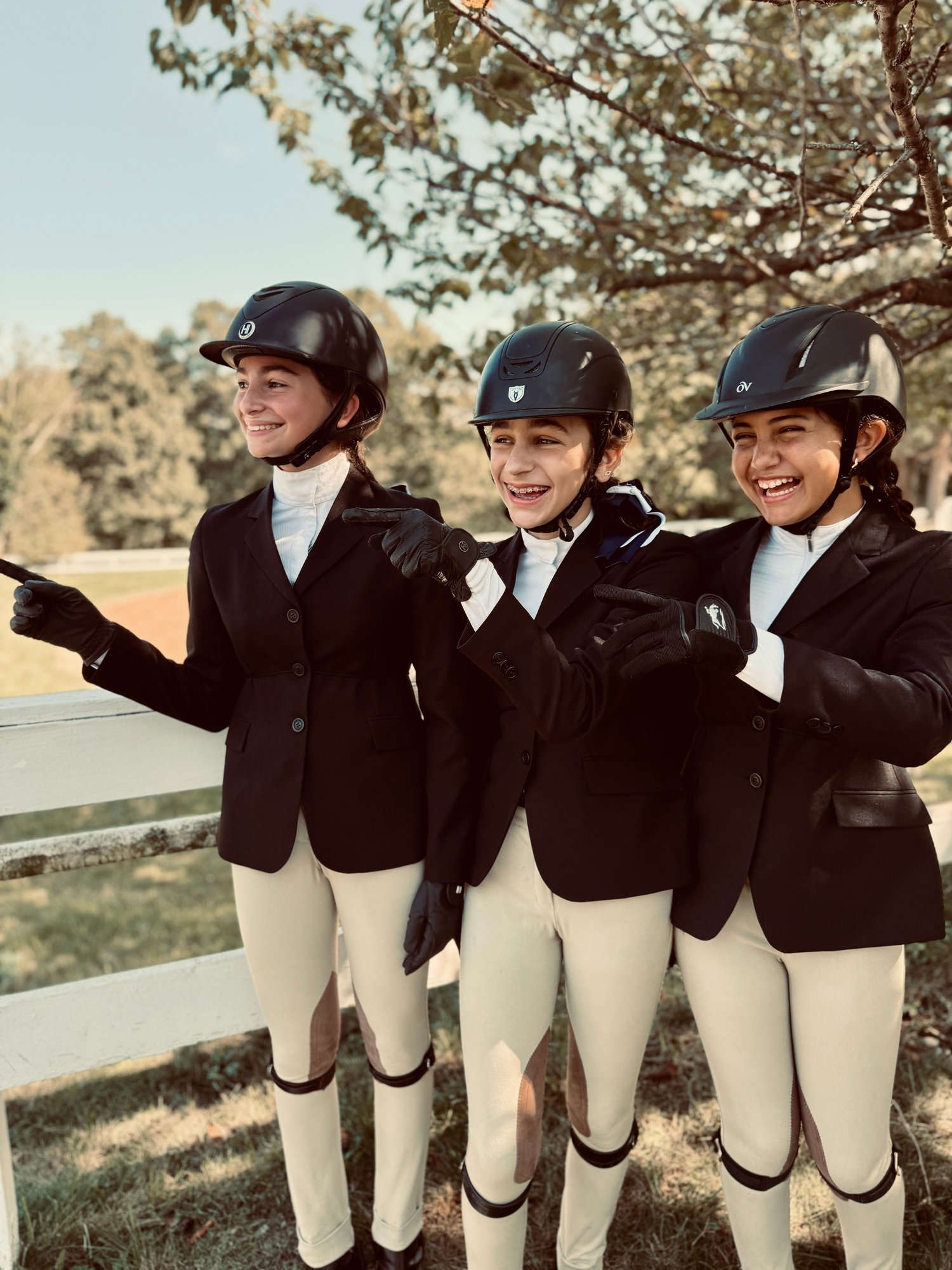 Three young girls in equestrian show attire—wearing black helmets, riding jackets, and beige breeches—smiling and pointing outdoors next to a white fence.
